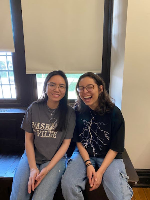 Two women sit on a radiator in front on windows with blinds down, one smiles while the other laughs