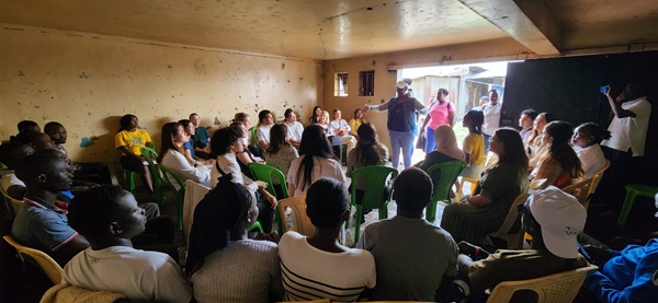 Many people sit in an arched formation in an underground room listening to a standing person speak